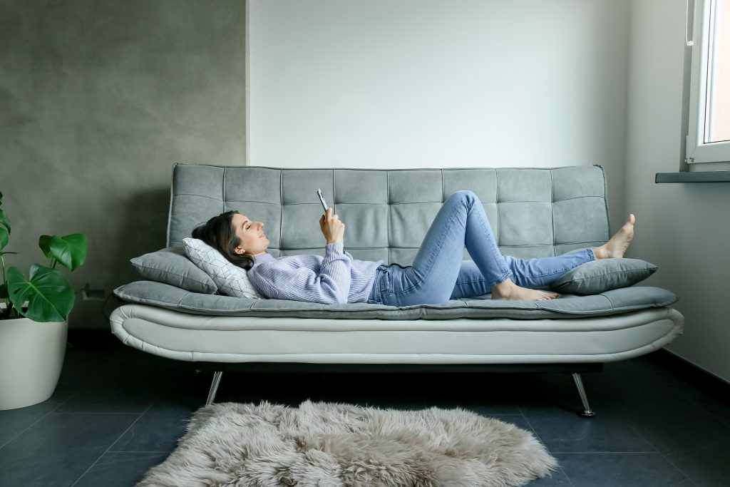 Back,View,Of,Happy,Young,Woman,Laying,On,Sofa,Checking